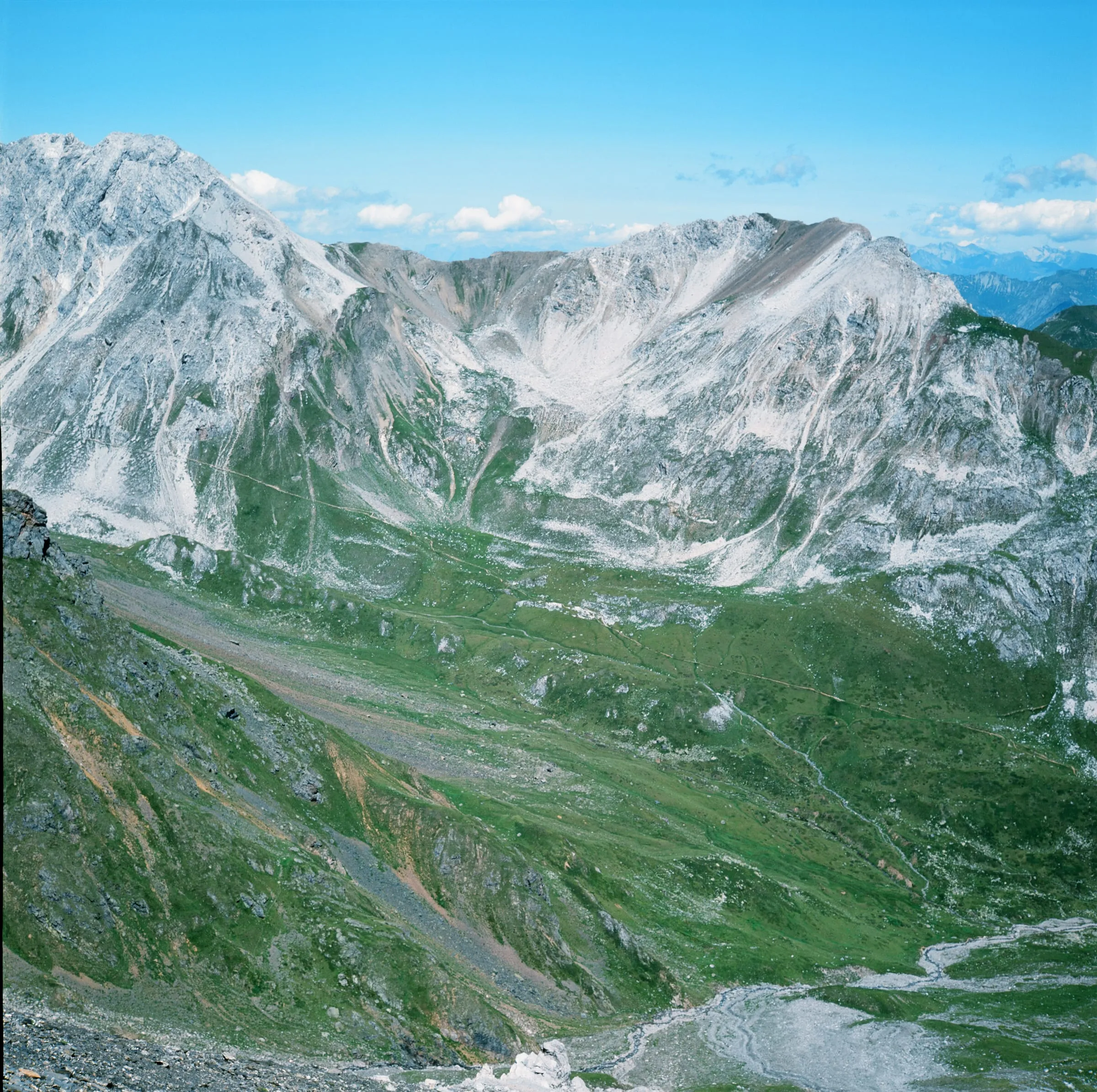 Farbaufnahme eines hochalpinen Talkessels: grüne Hänge unter hellen, zerklüfteten Kalkgipfeln, unten Geröll und ein Bergweg.
