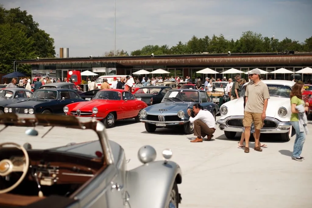 Parkplatz voller klassischer Sportwagen, Besucher begutachten die Fahrzeuge.