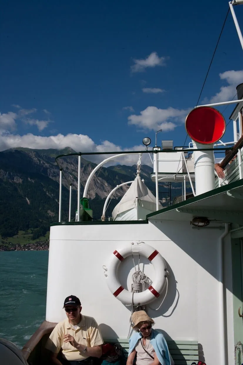 Passagiere sitzen an Deck eines Dampfschiffs neben einem Rettungsring, Bergkulisse im Hintergrund.